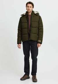 Jachetă puffer verde închis, cu fermoar frontal, design căptușit și glugă. Asortată cu un top maroniu, blugi negri și adidași maronii.