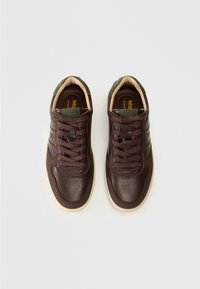 Sneaker in pelle marrone con suole color crema, caratterizzati da una punta perforata, dettagli cuciti e linguetta verde scuro. I lacci corrispondono al colore delle scarpe.