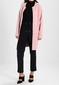 Nainen pukeutuu vaaleanpunaiseen auki olevaan neuletakkiin, mustaan ribbikuvioiseen poolopaitaan, mustiin kiristysnauhoilla varustettuihin housuihin ja mustiin paksupohjaisiin mokkasiineihin.