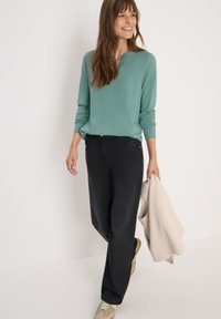 Un haut vert clair à manches longues avec un col henley, associé à un pantalon noir à jambes larges et des baskets beiges. Tient un pull de couleur crème.