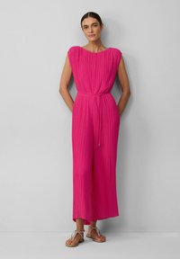 Ärmelloser, leuchtend pinker Jumpsuit mit plissierter Textur, tailliertem Schnitt und weitem Bein. Kombiniert mit flachen Sandalen mit Riemchen.