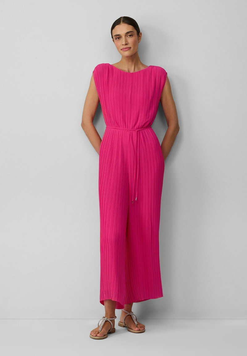 Ärmelloser, leuchtend pinker Jumpsuit mit plissierter Textur, tailliertem Schnitt und weitem Bein. Kombiniert mit flachen Sandalen mit Riemchen.