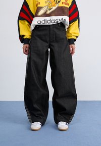 Denim preto de perna larga com riscas brancas laterais, combinado com um moletom gráfico colorido e ténis brancos com sola de borracha.