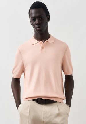 Νέος άνδρας με σκουρόχρωμο δέρμα και πλεξούδες φοράει ένα ανοιχτό ροζ πόλο μπλουζάκι με κοντά μανίκια, μπαίνει μέσα σε μπεζ παντελόνια με μαύρη ζώνη.