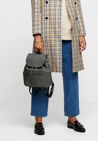 Sac à dos en cuir gris avec un rabat arrondi, une fermeture à cordon et une poche zippée à l'avant. Associé à un jean large bleu et un manteau à carreaux.