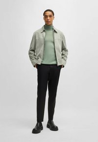 Gray suede jacket over a green turtleneck, black slim-fit trousers, and black ankle boots. The outfit features minimalistic design elements.