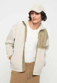 Chaqueta de forro polar para hombre en crema y oliva, con un cuello alto, bolsillo con cremallera y logo bordado. Llevada sobre una camiseta blanca.