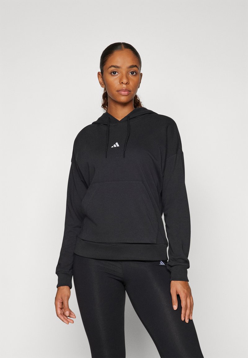 Sweatshirt preto com capuz, bolso frontal, cordões e logótipo branco no peito. Textura de tecido suave, ajuste relaxado, combinado com leggings pretas.