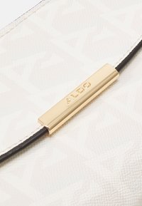 Bílá strukturovaná kabelka s jemným geometrickým vzorem, se zlatým logem a černým koženým popruhem.