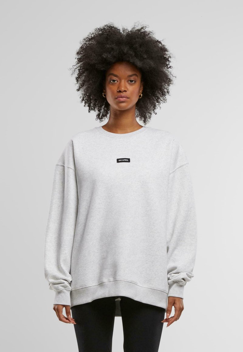 Sudadera gris claro de gran tamaño, fabricada en una tela suave, con cuello redondo, hombros caídos y una pequeña etiqueta negra con "OH APRIL" en el frente.