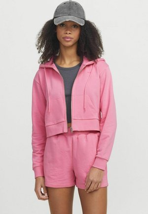 Felpa rosa tagliata con tasche frontali e zip, abbinata a pantaloni shorts rosa coordinati. Il modello indossa una canotta grigia e un cappellino grigio.