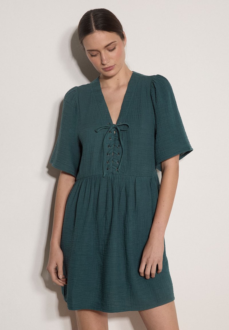 Frau mit zurückgebundenem dunklem Haar, die ein dunkelgrünes, strukturiertes Kleid mit kurzen Ärmeln und einer Schnürung am V-Ausschnitt trägt.