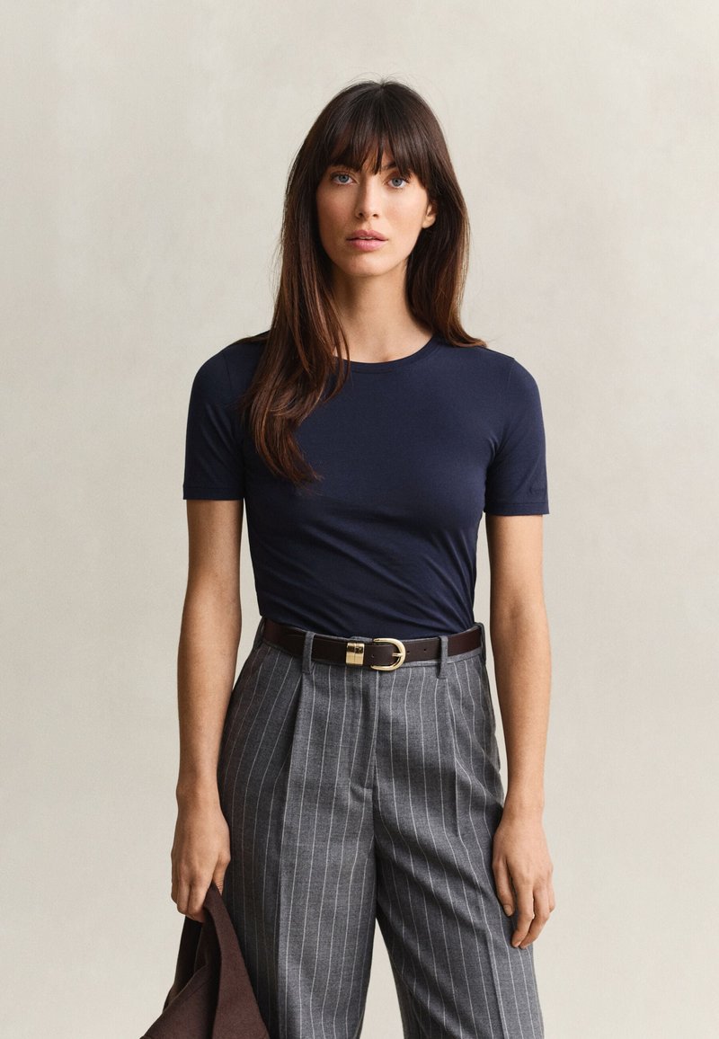 Femme aux longs cheveux bruns portant une chemise bleu marine à manches courtes et un pantalon gris à fines rayures, tenant une veste sombre sur un fond uni.