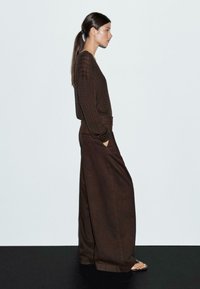 Bruine gebreide top met lange mouwen en een textuurpatroon, gecombineerd met chocoladebruine broek met wijde pijpen. Het model staat voor een lichte achtergrond.