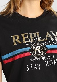 Close-up of a black T-shirt with gold "REPLAY" text, red and blue stripes, and white text saying "Love kills, you'll better stay home."