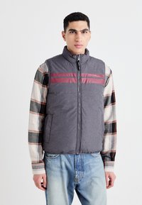 Chaleco gris sin mangas con cuello alto, presenta rayas rojas horizontales y un cierre frontal. Llevado sobre una camisa de cuadros de manga larga.