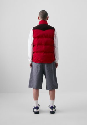 Red puffer vest with a brown collar, worn over a white long-sleeve shirt, paired with gray shorts and dark sneakers. Simple design.