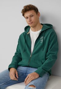 Grüner Zip-Hoodie mit Kordelzug und Kängurutaschen, über einem weißen T-Shirt getragen. Kombiniert mit hellblauen, verwaschenen Jeans.