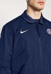 Laivaston tuulitakki, jossa on vetoketju, taitettava kaulus, valkoinen Nike-logo vasemmassa rinnassa ja Paris Saint-Germainin vaakuna oikealla.