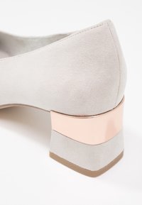 Chaussure en daim gris clair avec un bout arrondi, dotée d'un petit talon carré, accentuée par une bande métallique dorée rose.