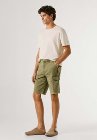 Young man with curly hair stands, wearing beige t-shirt, olive green cargo shorts, and tan slip-on shoes against plain white background.