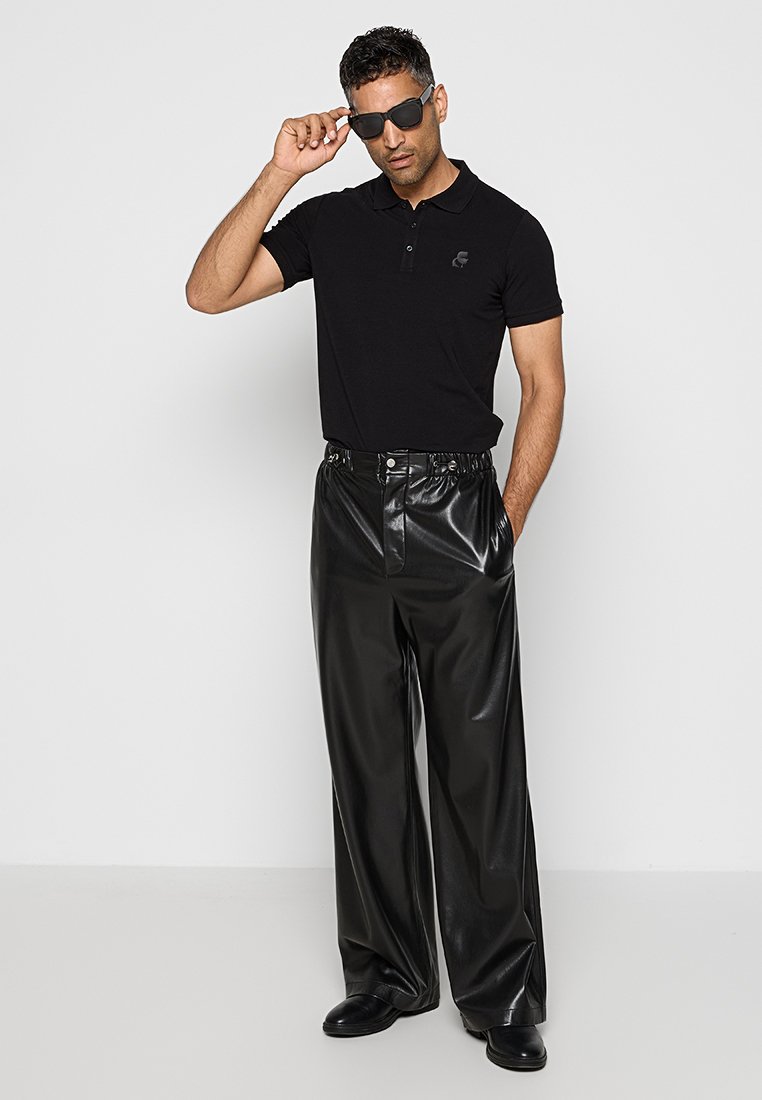 Polo noir avec un logo discret, associé à un pantalon en cuir noir brillant taille haute et des chaussures noires. Accessoirisé avec des lunettes de soleil foncées.