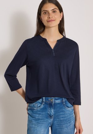 Femme aux longs cheveux bruns portant un top bleu marine à manches trois-quarts et un jean bleu, debout avec une main sur la hanche.