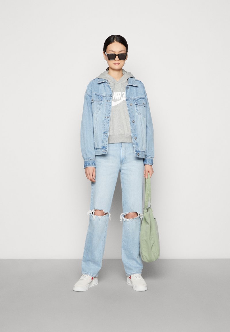 Světle modrá džínová bunda, šedá mikina s logem, světle modré džíny s trhlinami, bílé tenisky a zelená taška přes rameno. Černé sluneční brýle.