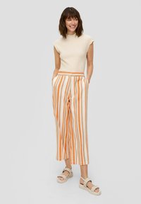 Lichtbeige gebreide top gecombineerd met wijde broek met verticale oranje strepen. Het model draagt gestructureerde sandalen.
