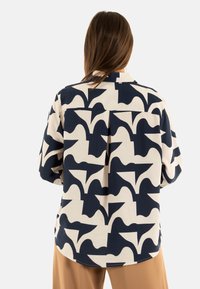 Camisa de manga larga confeccionada con una tela que presenta un patrón abstracto en tonos azul marino y crema. La parte posterior cuenta con un plisado central y un cuello clásico.