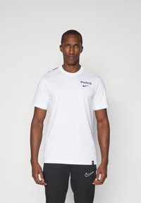 Hvid bomulds t-shirt med "PARIS" i marineblå tryk, Nike-logo på brystet og sorte detaljer på skuldrene. Korte ærmer, rund hals.