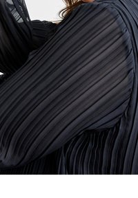 Černá skládací textilie s jemnou strukturou, s dlouhými, volně splývajícími rukávy, které se elegantně vlní. Detailní vertikální sklady po celé ploše.