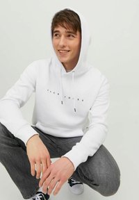 Un sweat à capuche blanc avec un logo sur le devant, une capuche ample à cordon, des manches resserrées, associé à un jean gris et des chaussures noires, sur un fond neutre.