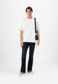 Young man standing wearing a white t-shirt, dark blue jeans, gray sneakers, and a black shoulder bag against a white background.