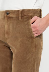 Καφέ βελούδινες παντελόνες με ευθεία κοπή, διαθέτοντας διαγώνιες μπροστινές τσέπες και κλείσιμο με κουμπί στη μέση. Απαλή υφή.