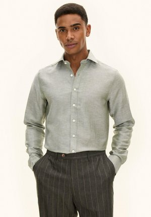 Mann trägt hellgraues Hemd mit Knöpfen und dunkle Nadelstreifenhose, Hände in den Taschen, steht vor einfachem hellem Hintergrund.