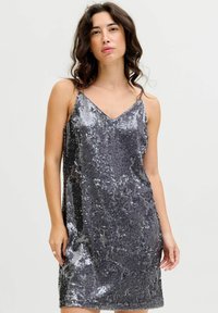 Srebrna sukienka z cekinami, z dekoltem w kształcie litery V, cienkimi ramiączkami i luźnym krojem. Powierzchnia ma refleksyjne, teksturowane wykończenie z różnymi wzorami cekinów.