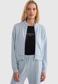 Gaiši zila rāvējslēdzēja jaka no mīksta auduma, ar augstu apkakli un ribotām piedurknēm. Valkāta virs melnas krekla ar baltu tekstu. Brīva piegriezuma bikses.