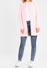 Cardigan lungo rosa chiaro senza colletto, abbinato a jeans skinny blu con strisce scure sui lati e sneakers bianche.