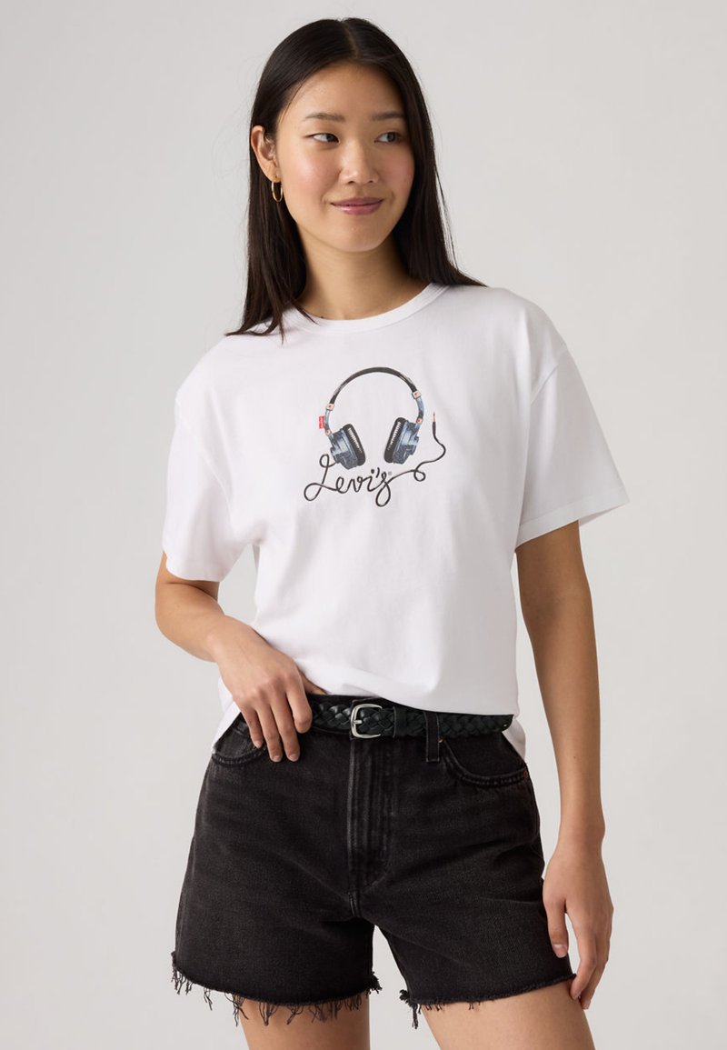 Νεαρή γυναίκα που φοράει λευκό T-shirt με γραφικό ακουστικών και το κείμενο «Levi's», συνδυασμένο με μαύρο τζην σορτς και πλεξούδα ζώνη.