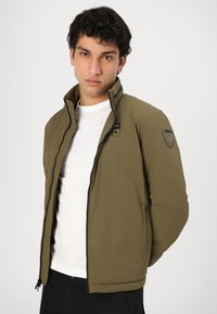 Veste bomber vert olive avec fermeture éclair, col haut et surface texturée ; présente un patch logo sur l'épaule et des poches latérales.