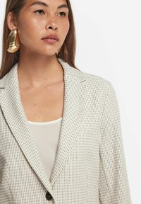 Randig beige kavaj med slag och en enkel knappstängning, textil med struktur, i kombination med en vit topp och stora gyllene örhängen.