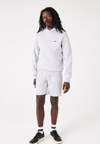 Completo di felpa grigia e pantaloni corti realizzato in tessuto morbido, con un logo verde. Il modello indossa sneakers nere e calzini a fantasia.