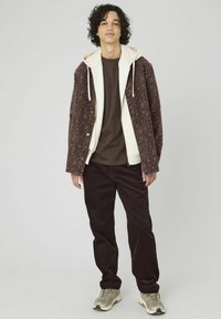 Veste marron à motifs avec doublure crème, chemise marron et pantalon en velours côtelé foncé. Portée avec des baskets grises. Cheveux bouclés.