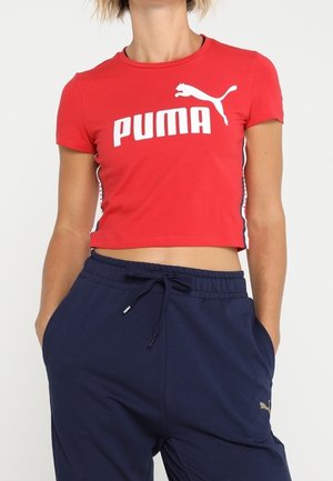 Person iført en rød cropped Puma t-shirt og marineblå Puma sweatpants med hænderne i lommerne, stående mod en ensfarvet baggrund.