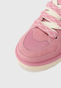 Gros plan sur l'avant d'une basket rose avec des textures en suède et mesh, présentant des lacets roses et blancs à motifs sur un fond clair.