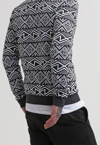 Man i en svart och vit geometrisk mönstrad långärmad skjorta med vit t-shirt under och svarta byxor, som står mot en enfärgad bakgrund.