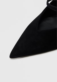 Μαύρο σουέντ παπούτσι με μυτερή μύτη και σχέδιο σε σχήμα V, με λεπτό λουρί στον αστράγαλο. Λείο υφή και μινιμαλιστικό στυλ.