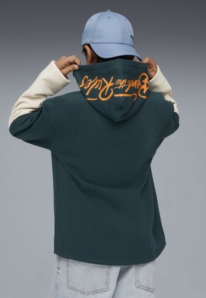 Sudadera con capucha en verde oscuro con mangas de crema, que presenta texto naranja en la parte posterior. El material parece suave y tiene un ajuste relajado. Se lleva una gorra azul claro.