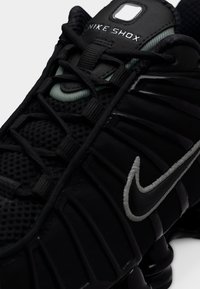 Zapatilla Nike Shox negra con parte superior de malla texturizada, detalles reflectantes, sistema de cordones prominente y el logo característico de la marca.